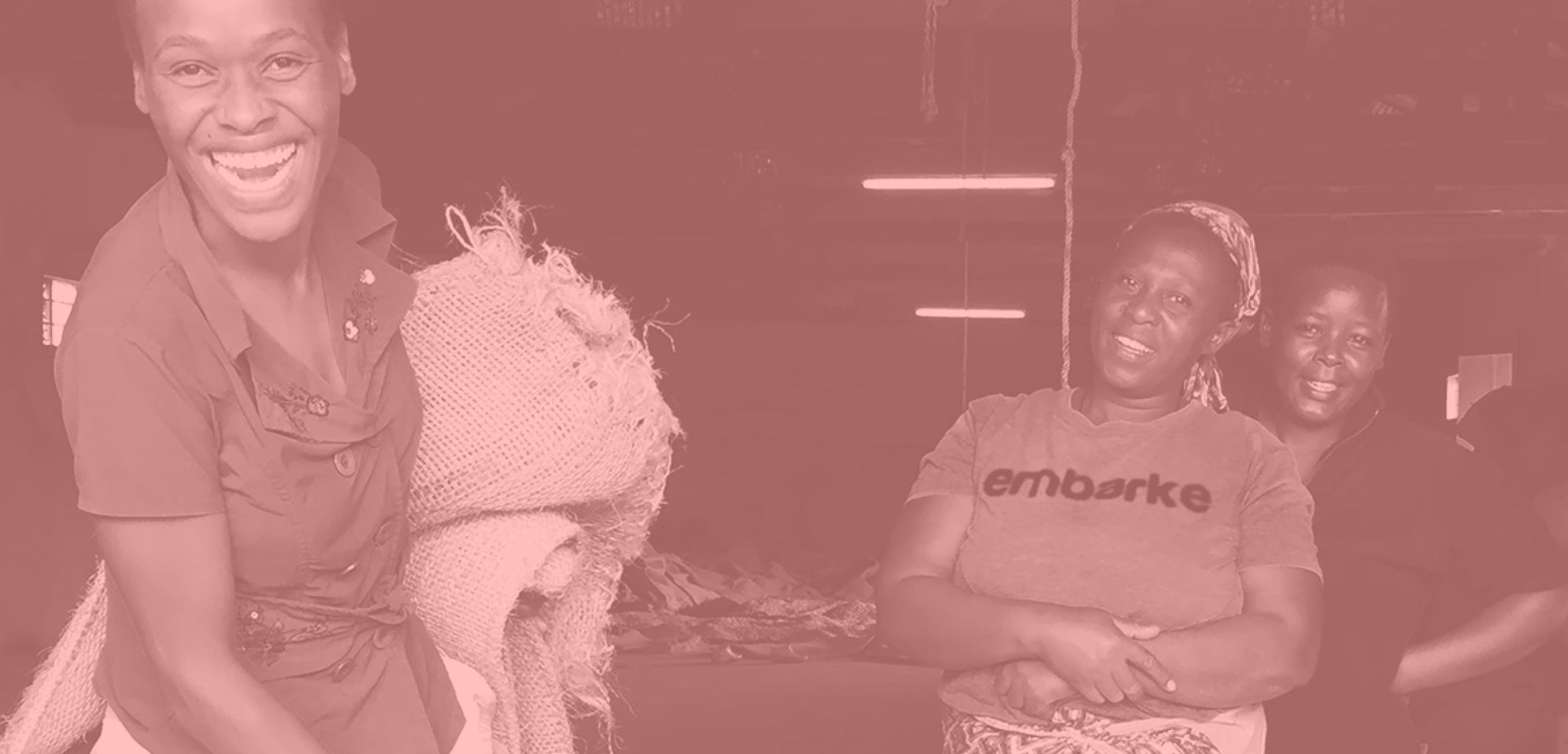Three women smiling indoors; one holds a large burlap sack, and another wears a shirt that says "embarke".