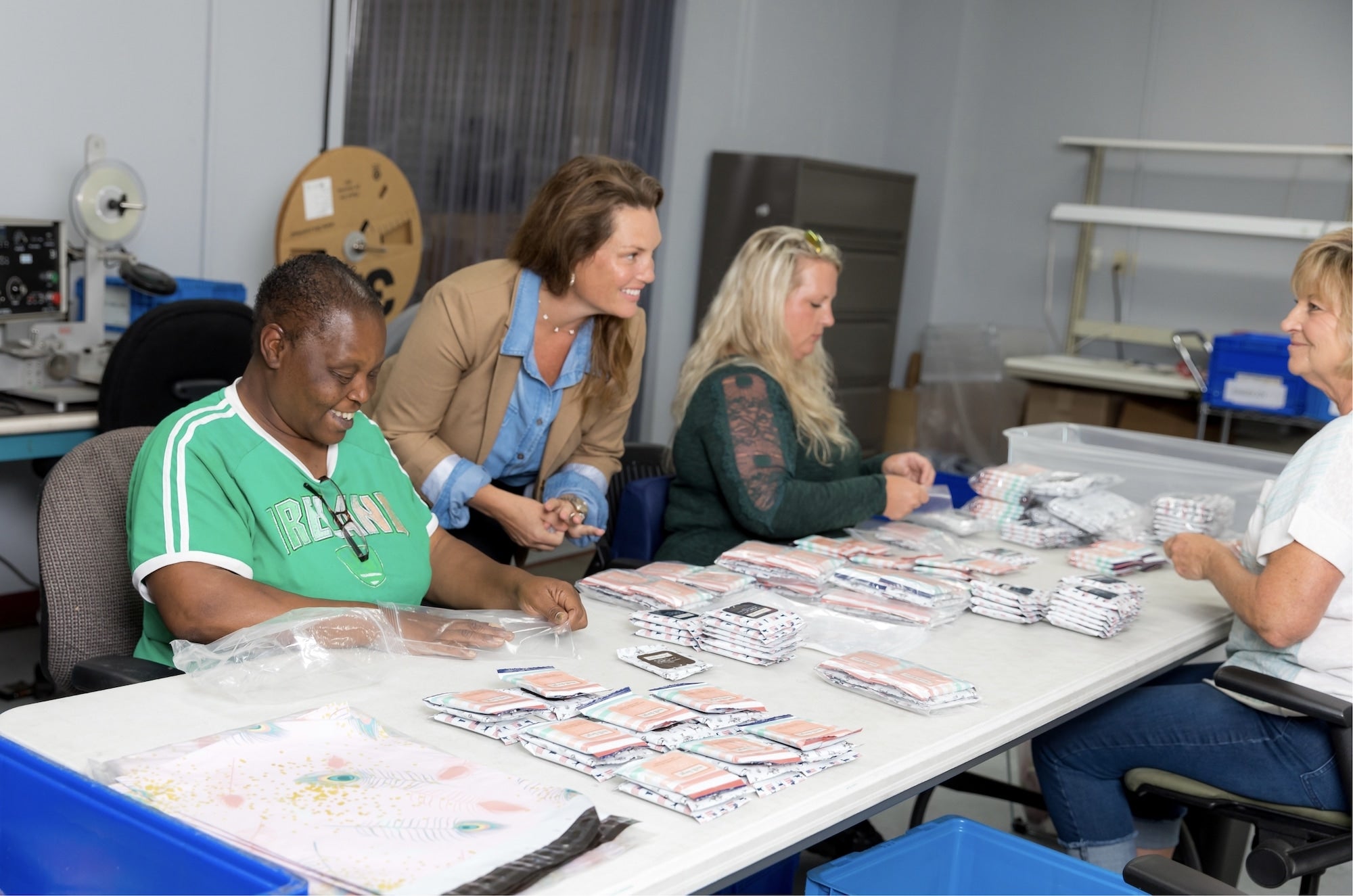 Four women sit and stand around a table, sorting and packaging folded fabric items in a workspace.