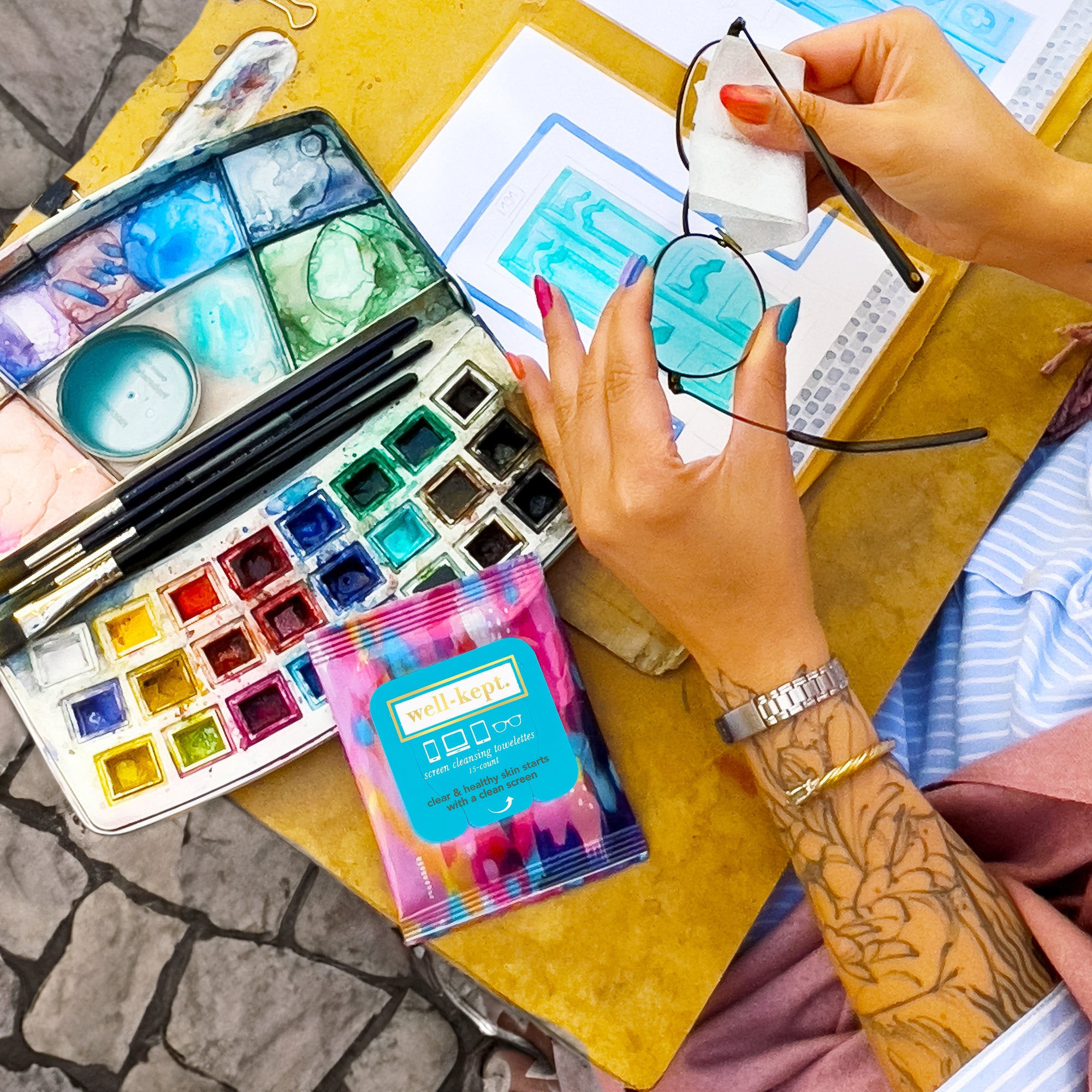 A person cleaning glasses with a wipe beside a watercolor palette and art supplies on a yellow table.