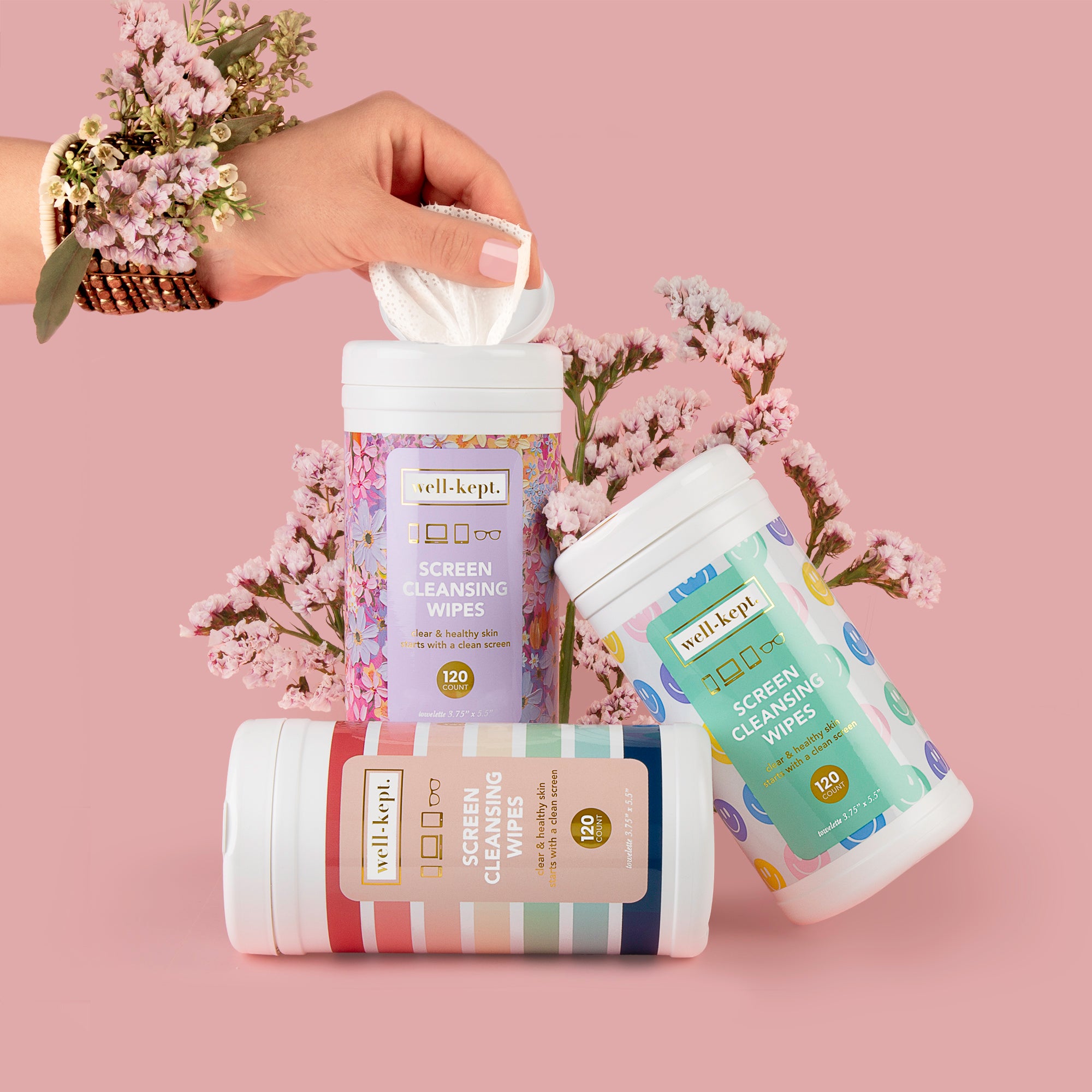 A hand pulls a pre-moistened towelette from the NEW SIZE - CANISTER TRIO screen cleansing wipes container, with flowers in the background.