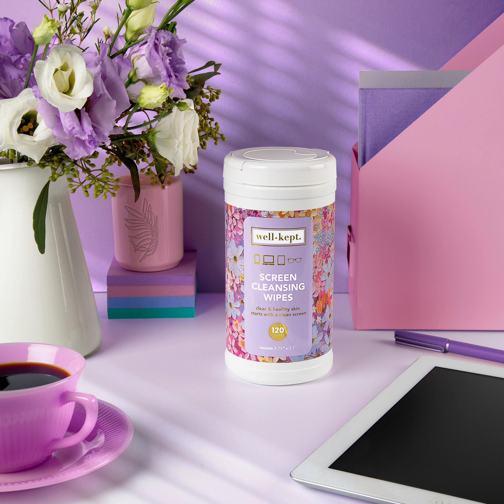 The NEW SIZE - FLOWER POWER CANISTER of pre-moistened towelettes sits on a desk with flowers, a cup, notebooks, and a tablet—perfect for tackling tech germs and keeping electronics spotless.