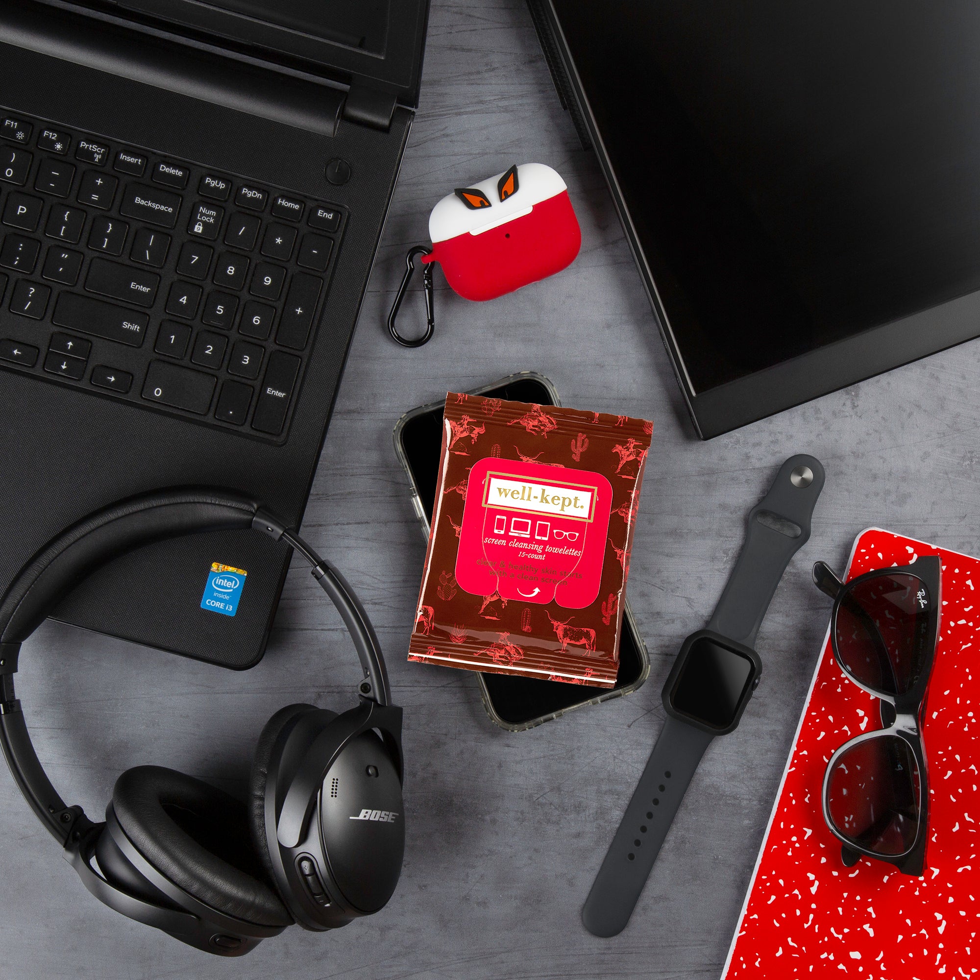 Flat lay of the TEXAS HOLD, 'EM laptop, headphones, smartwatch, sunglasses, AirPods, electronics cleaning wipes, and a red notebook displayed on a desk.