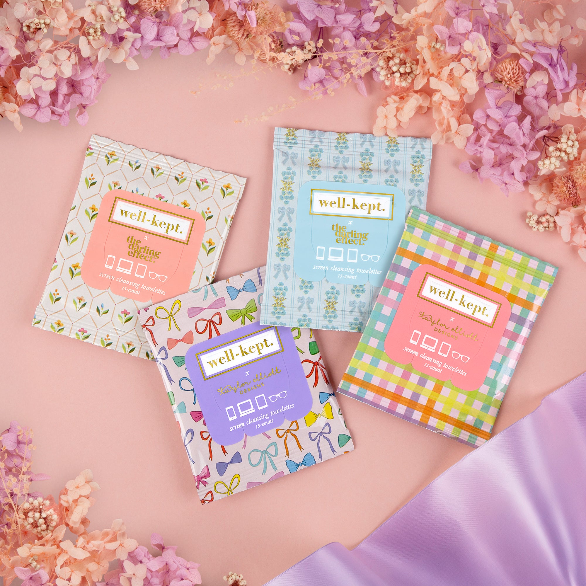 Four colorful Well-Kept screen cleaning wipes surrounded by pink flowers on a pastel background.