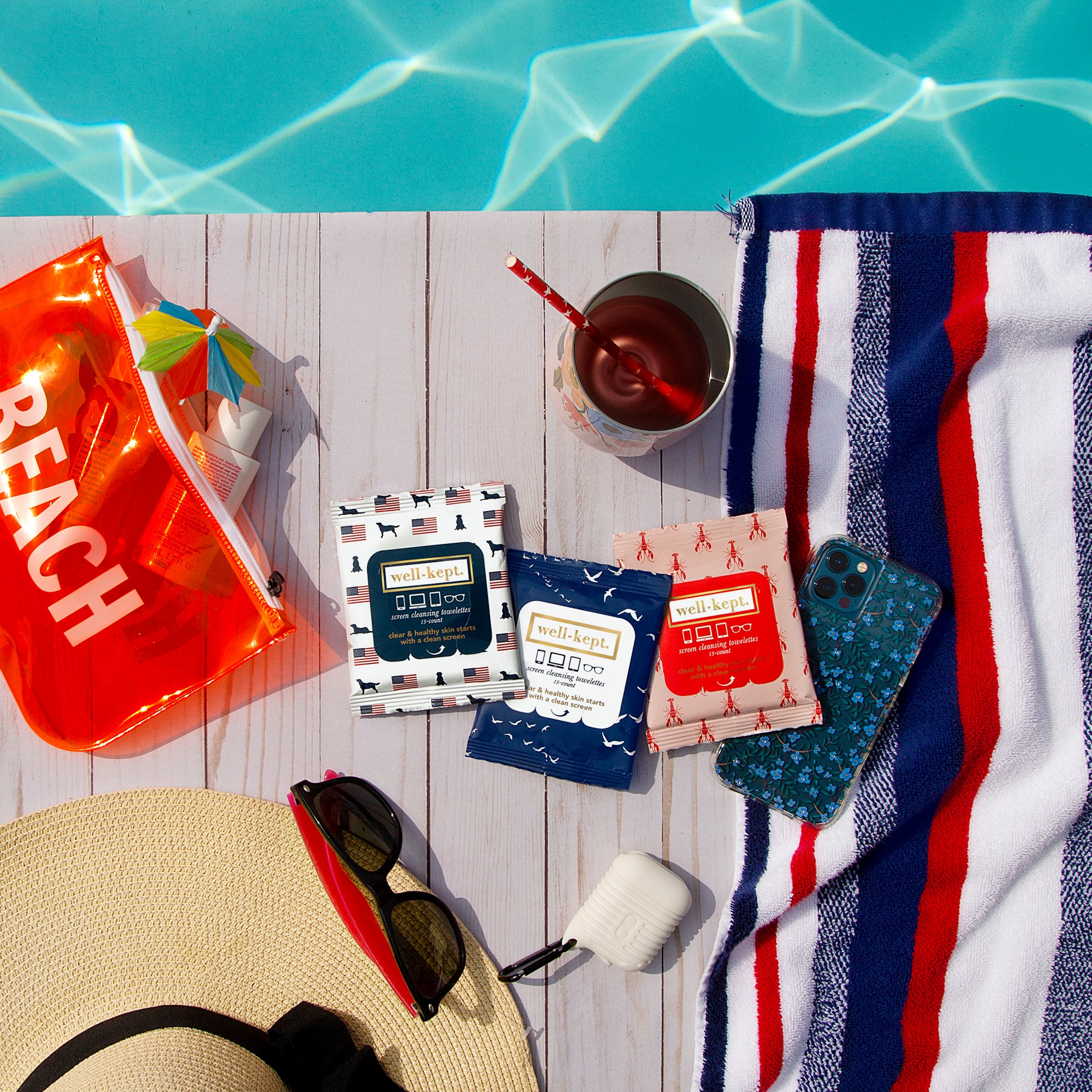 Flat lay of poolside essentials: towel, phone, sunglasses, drink, hat, and a LOBSTERFEST - TWELVE PACK (12) of pre-moistened towelettes—perfect for electronics cleaning while you relax in style.