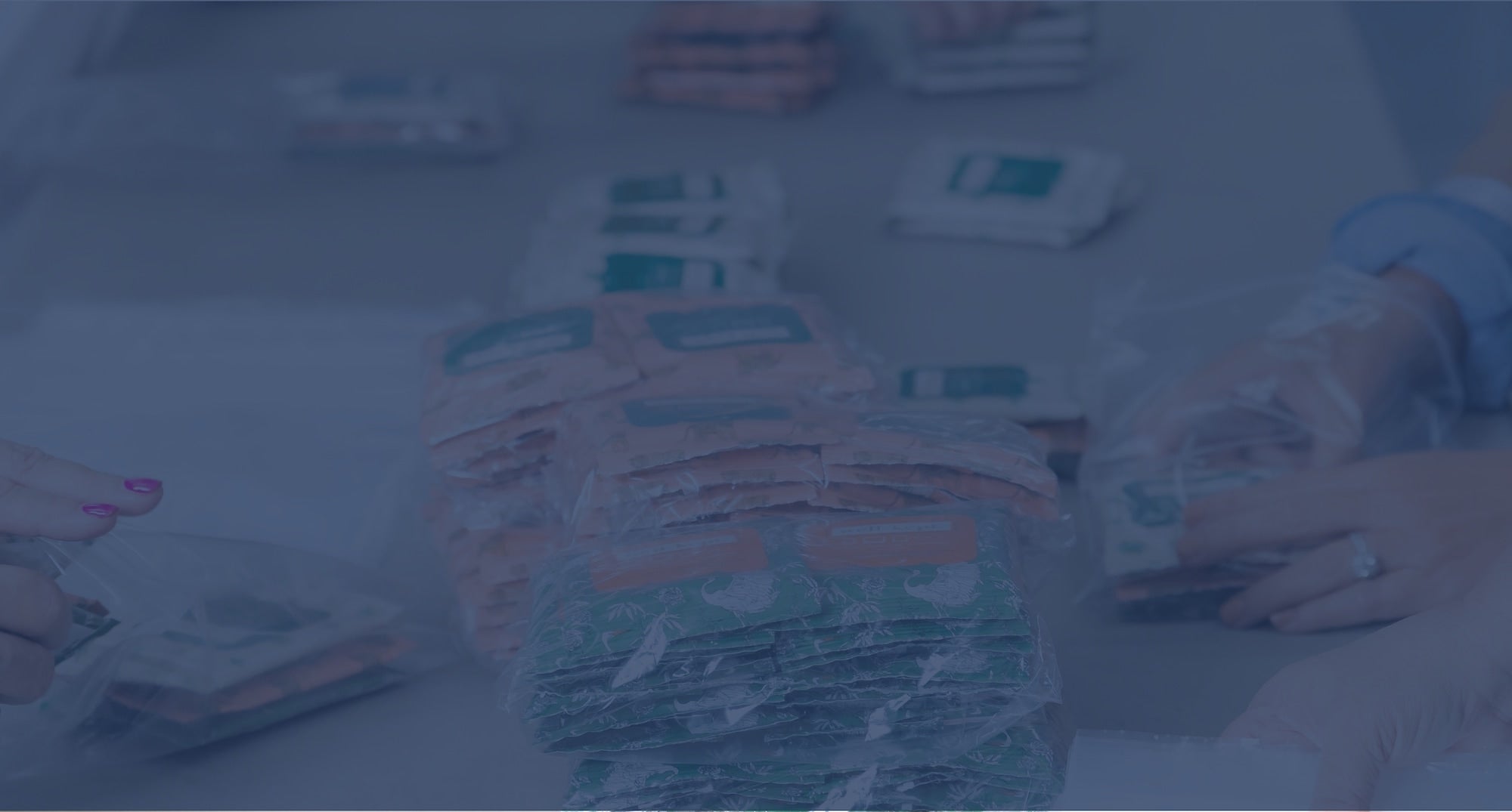 Three people pack stacks of assorted food pouches into clear plastic bags on a table.