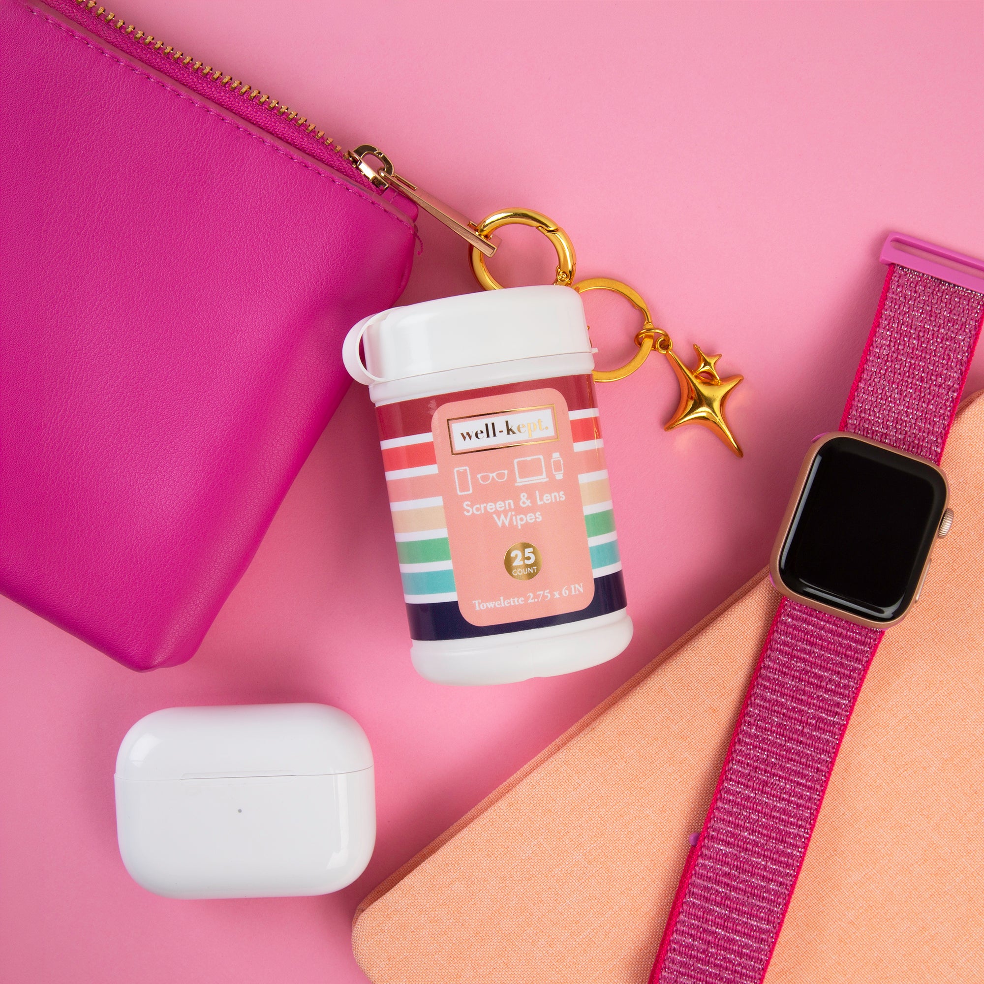 A flat-lay of a well-kind Screen & Lens Wipes container with a gold star charm, alongside a white charging case, a pink zip pouch, a peach notebook, and a pink smartwatch band on a pink background.