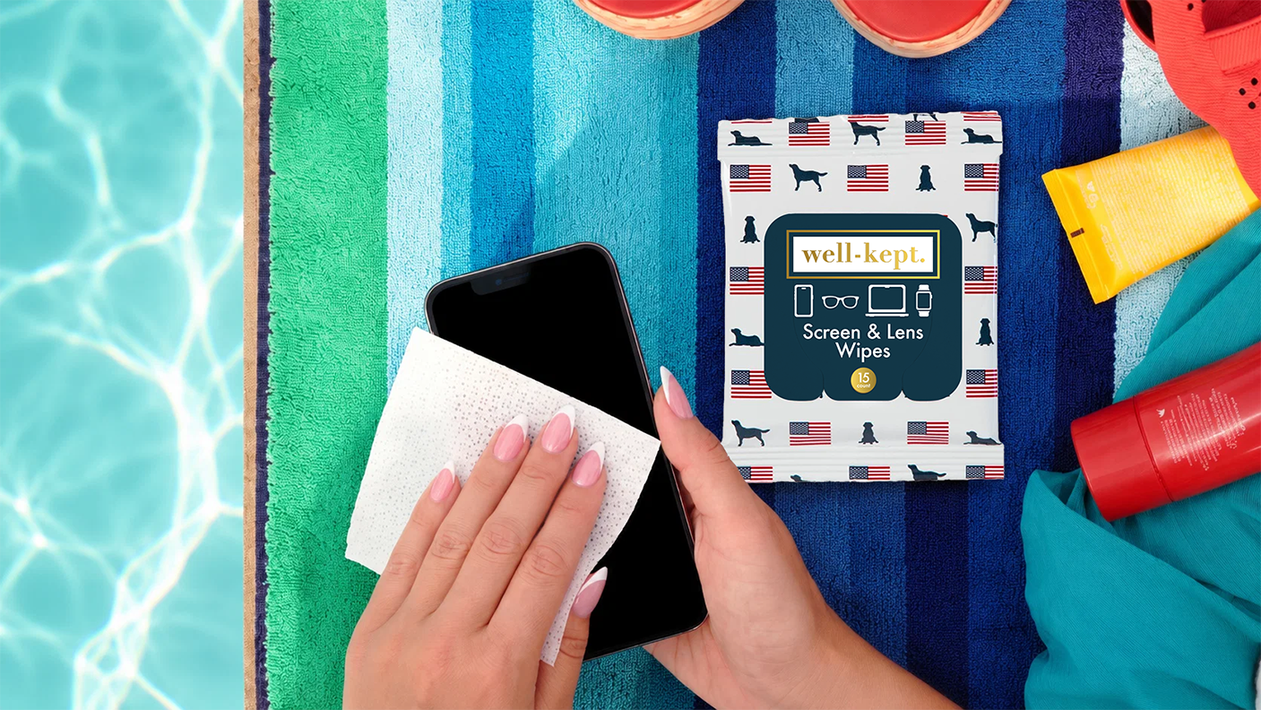 Hand cleaning a phone screen with a wipe next to an Old Glory flag and dog print Well-Kept pack, on a striped towel by a pool
