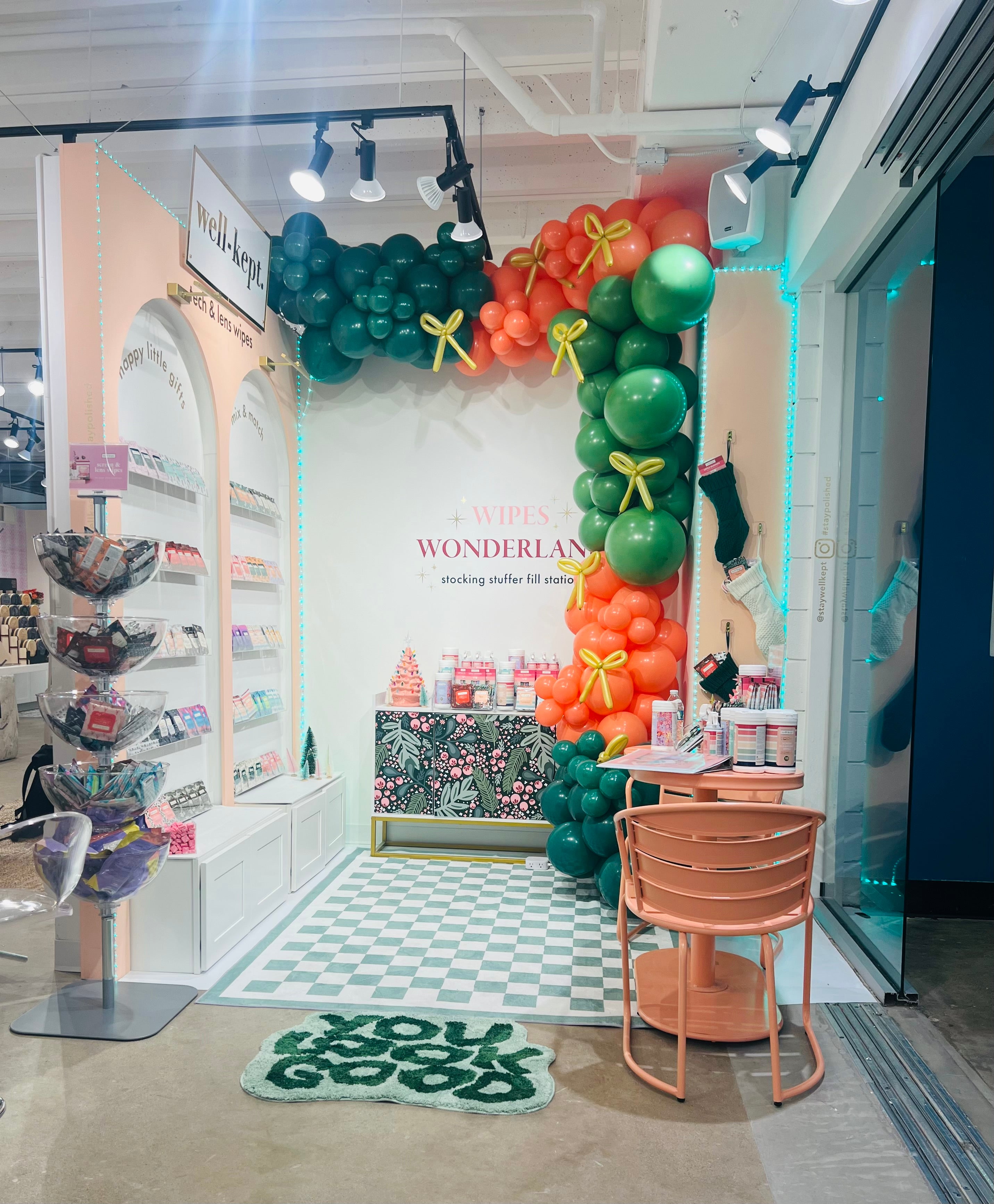 Festive retail setup with colorful balloons, shelves of products, a green "YOU GO GIRL" mat, and the ACRYLIC 5-BOWL FLOOR DISPLAY for organized wipes in clear acrylic bowls.
