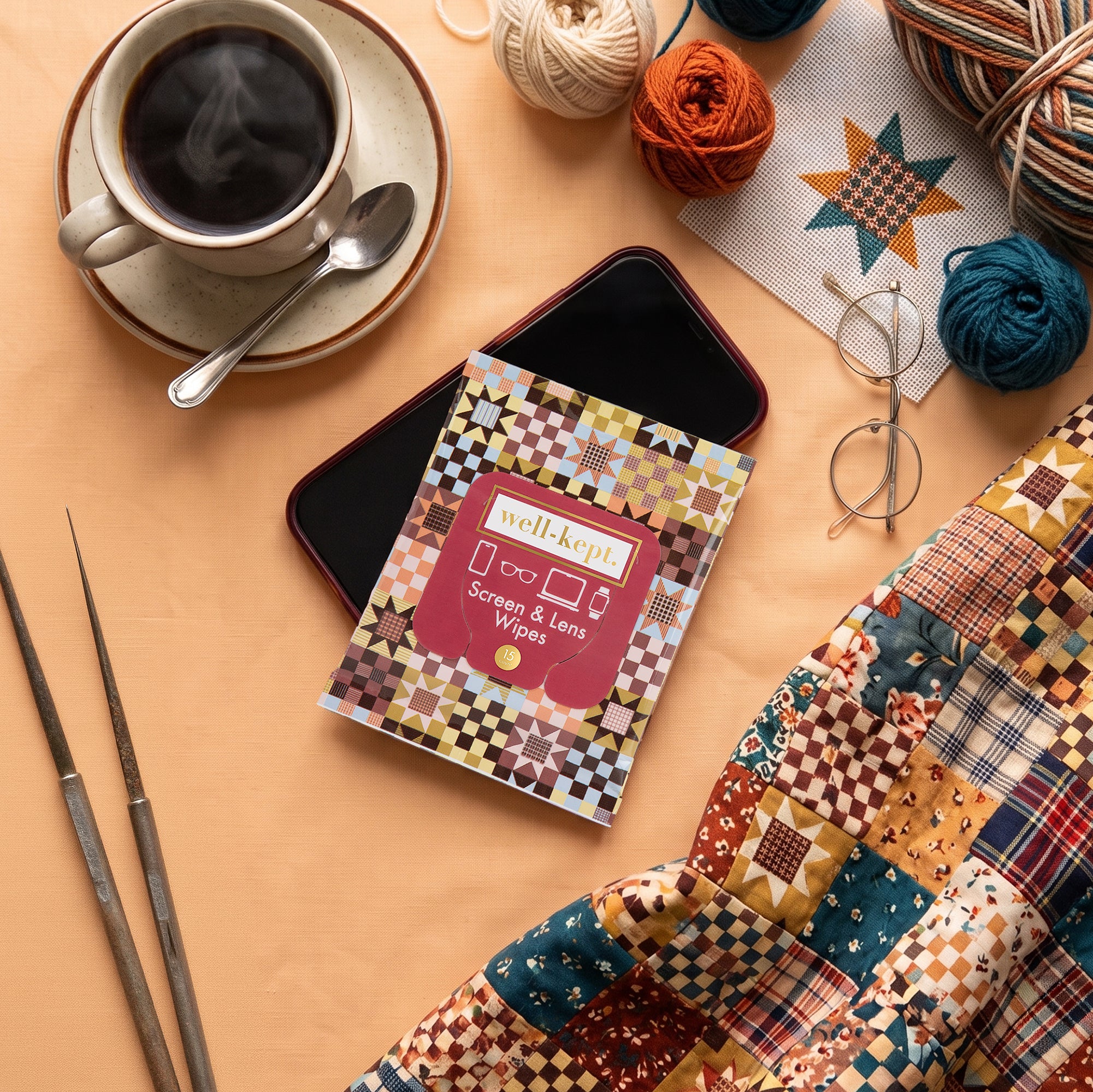 Storyteller quilt-pattern Well-Kept screen and lens wipes pack styled with a cup of coffee, glasses, yarn balls, knitting needles, and a quilt