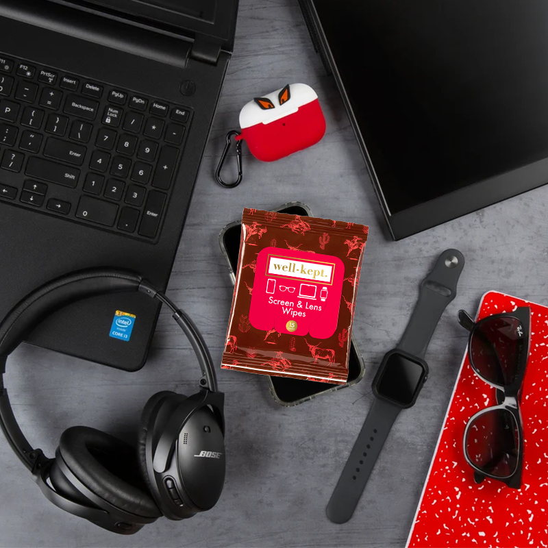 Texas Hold 'Em red print Well-Kept screen and lens wipes pack styled with headphones, a laptop, a smartwatch, and sunglasses on a desk