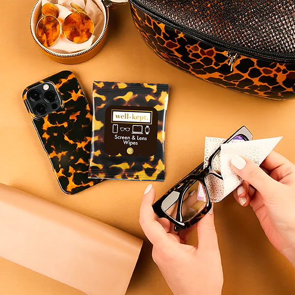 Tortoiseshell print Well-Kept screen and lens wipes pack with a hand cleaning glasses, next to a phone case and jewelry tray