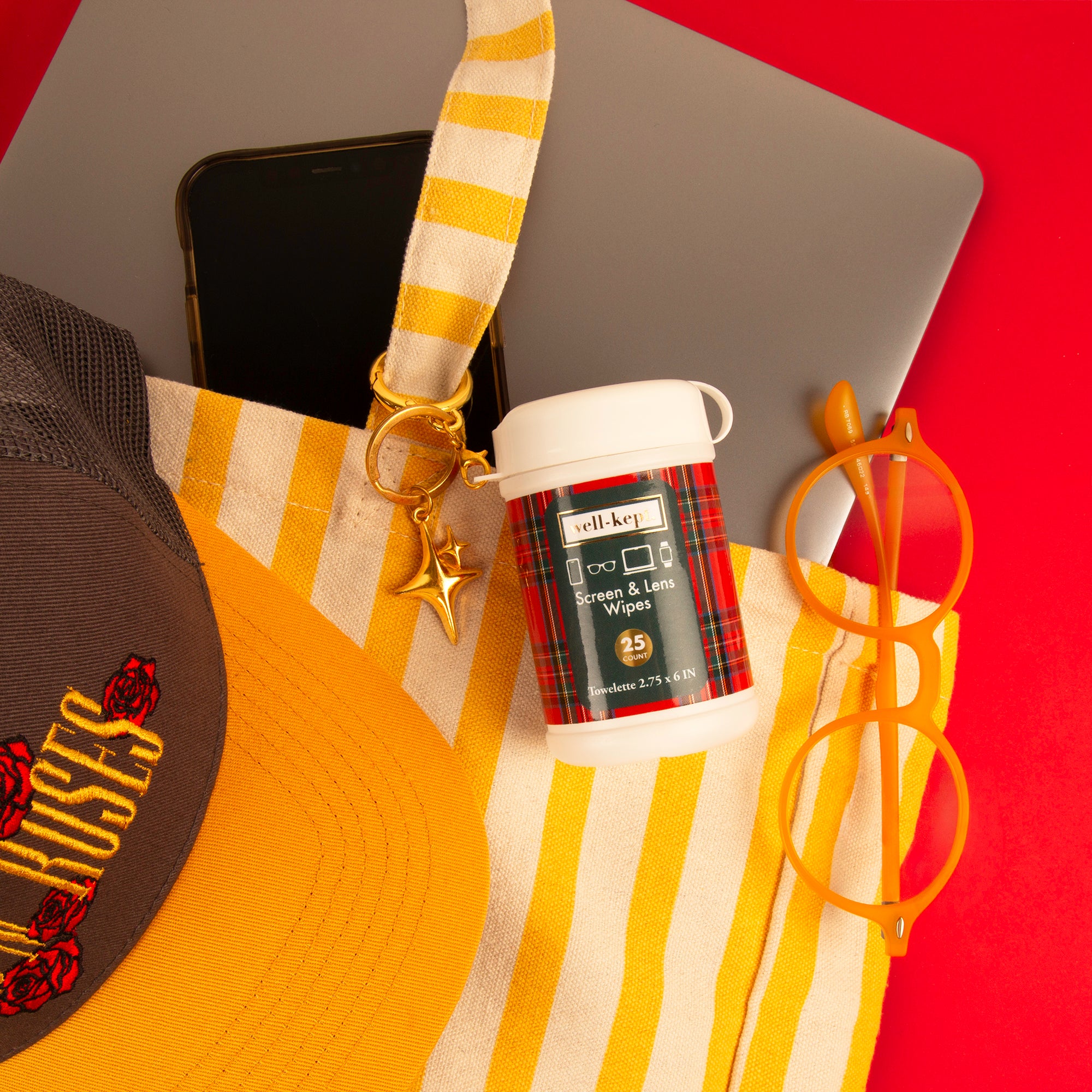 A small white plastic bottle of Wilky's Hand Sanitizer & Gel Wipes with a red plaid label, styled in a flat-lay with a yellow and white striped tote bag, a laptop, a phone, orange glasses, and a baseball cap on a bright red background.