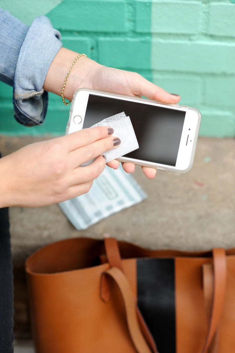 Hand cleaning a phone screen with a Well-Kept wipe, next to a leather tote bag against a teal and white brick wall