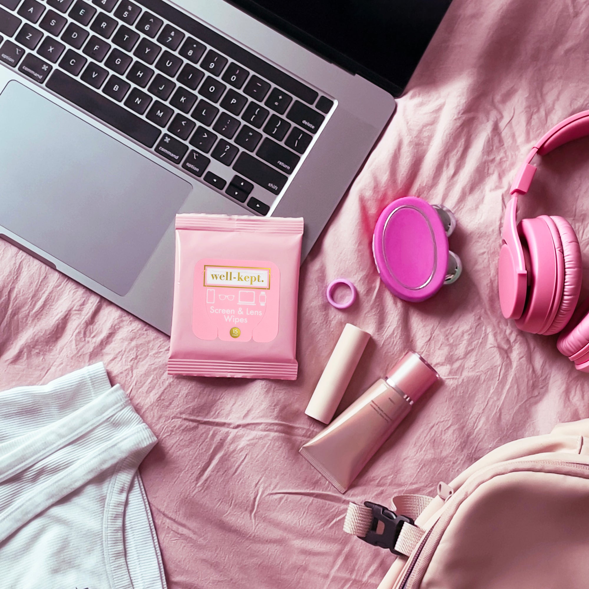 Pink Well-Kept screen and lens wipes pack styled with a laptop, headphones, a compact mirror, and a backpack on pink bedding