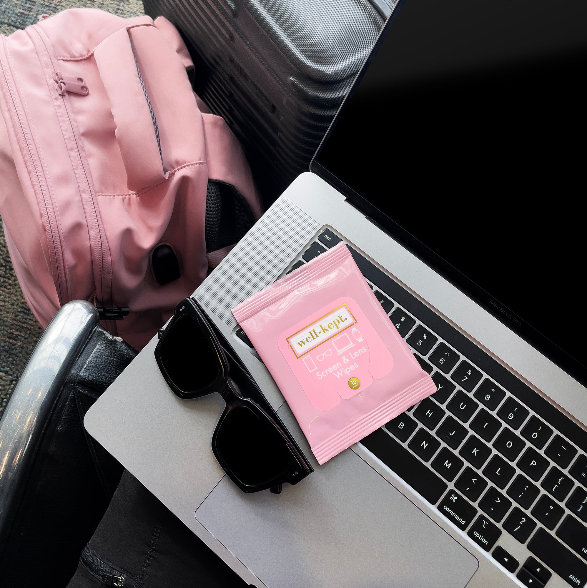 Pink Well-Kept screen and lens wipes pack resting on a laptop keyboard next to sunglasses and a pink backpack