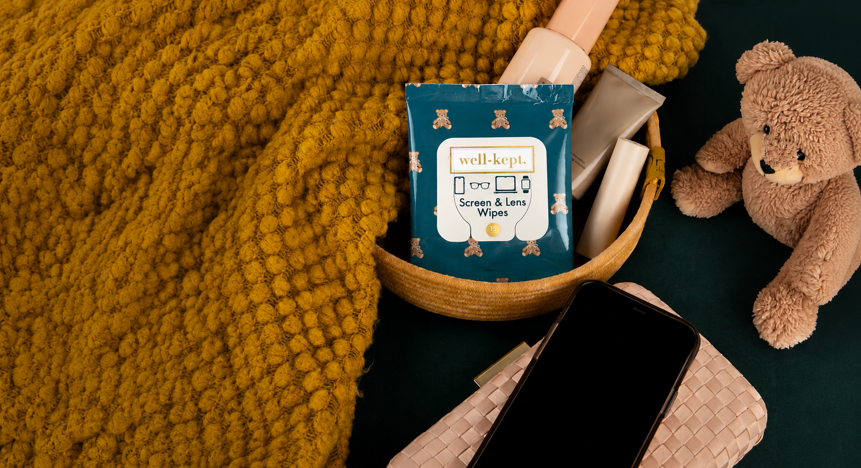 A flat-lay scene featuring a package of well-kept Screen & Lens Wipes in a small wooden basket, surrounded by cozy props including a chunky mustard yellow knit blanket, a brown teddy bear, a smartphone, and a woven clutch on a dark green surface.
